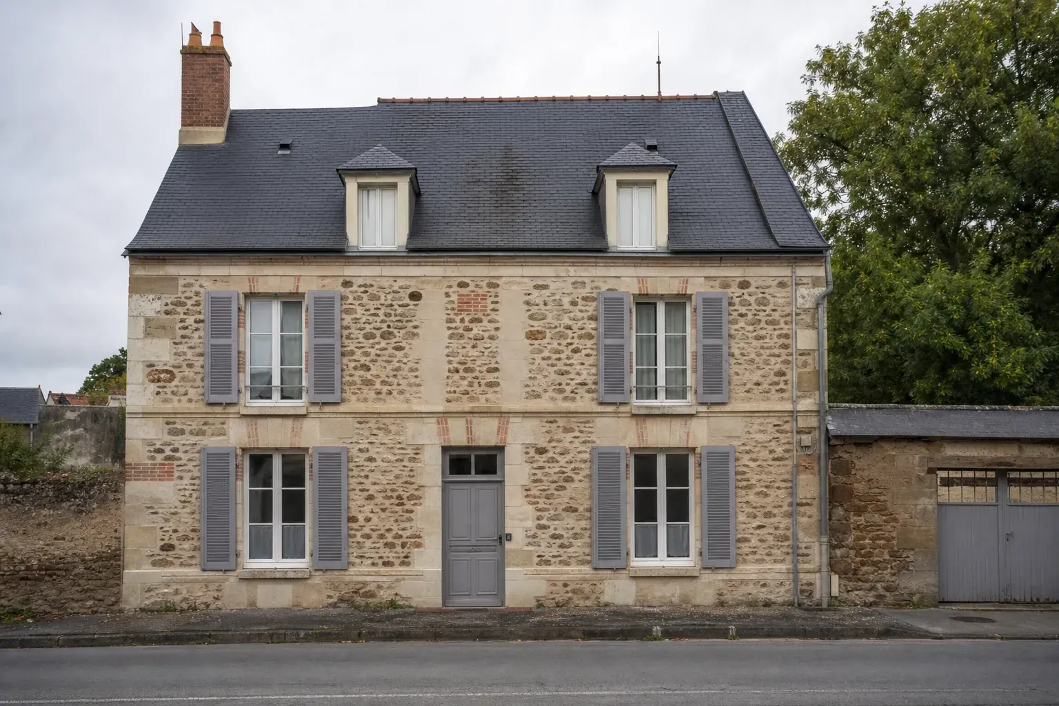 Maison de bourg sarthoise vue de face, toit d’ardoise à forte pente, deux lucarnes et une haute souche de brique