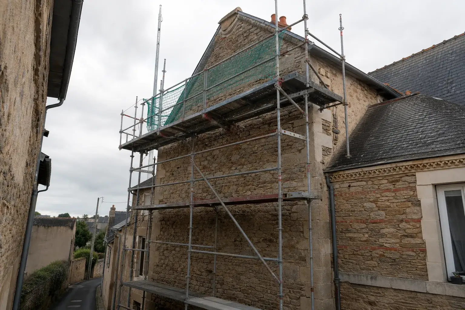 Échafaudage tubulaire monté contre le pignon d’une maison de pierre, dans une rue étroite qui limite la place de travail