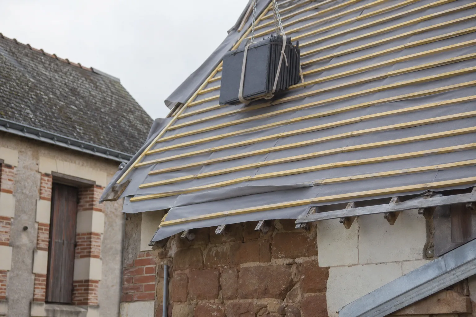Versant mis à nu, liteaux neufs posés sur un écran de sous-toiture gris, paquet d’ardoises hissé sur les liteaux