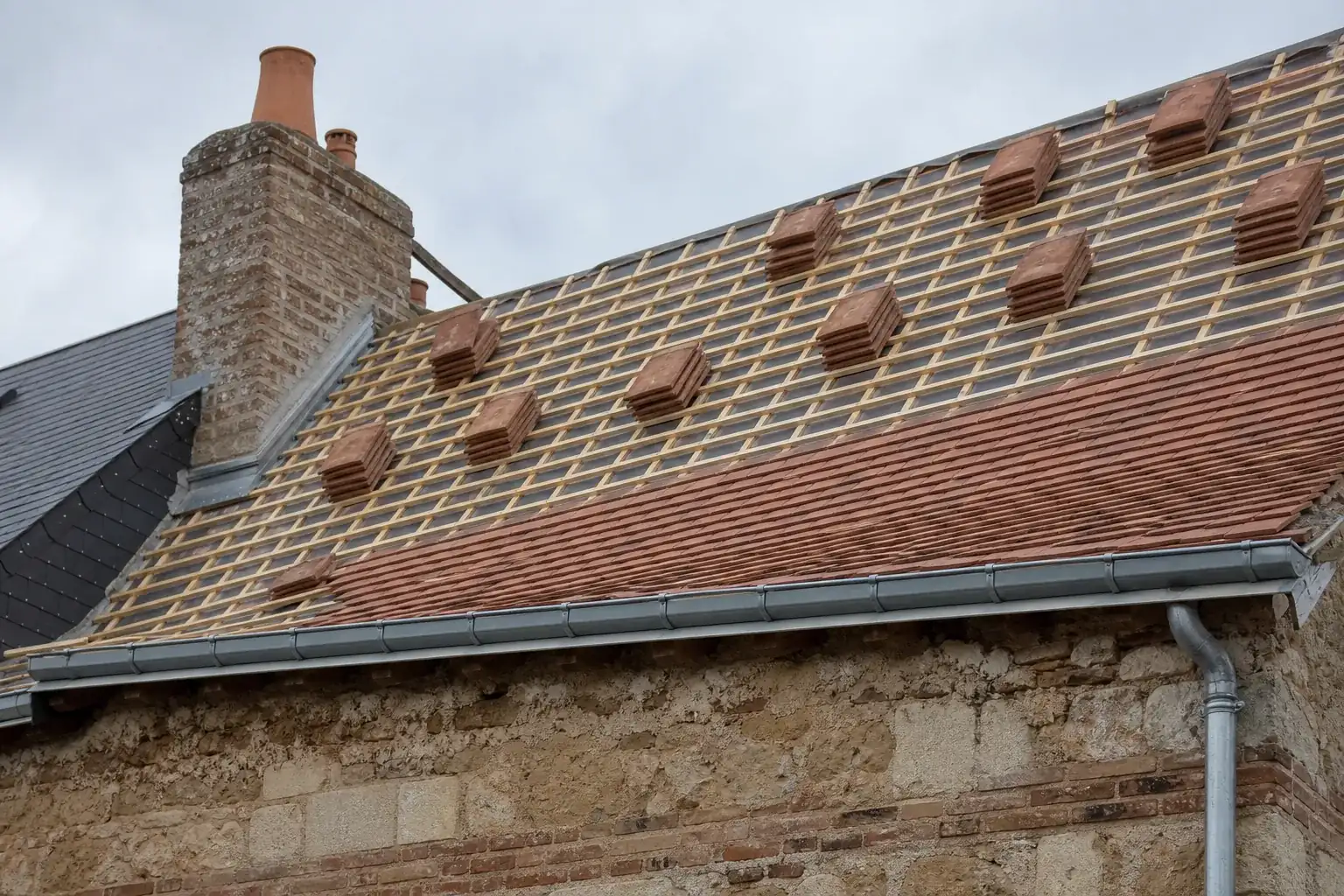 Versant de petites tuiles plates en cours de pose, accrochées rang par rang, piles réparties le long de la pente